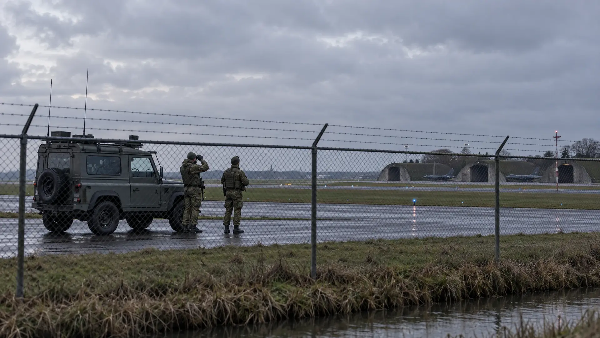 Twee militairen in camouflagekleding kijken op een betonnen platform langs een hekwerk naar een lege grijze lucht, met een patrouillevoertuig en lage hangars van een Nederlandse militaire vliegbasis op de achtergrond